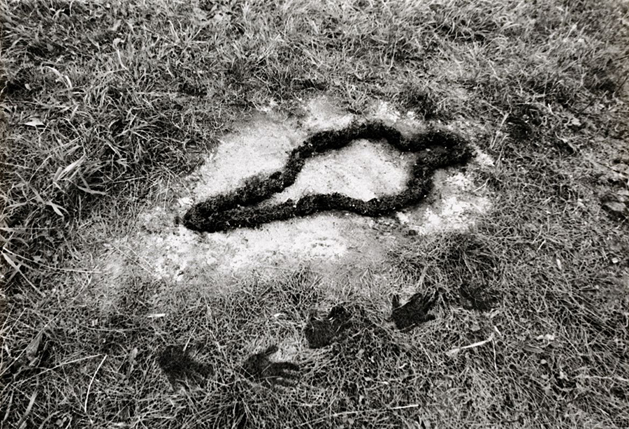 Silueta de un cuerpo femenino excavada sobre el pasto y delineada con tierra oscura, con huellas de manos en la parte inferior.