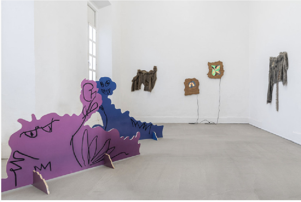 Sala blanca con siluetas de madera pintadas en morado y azul, y piezas textiles en la pared.