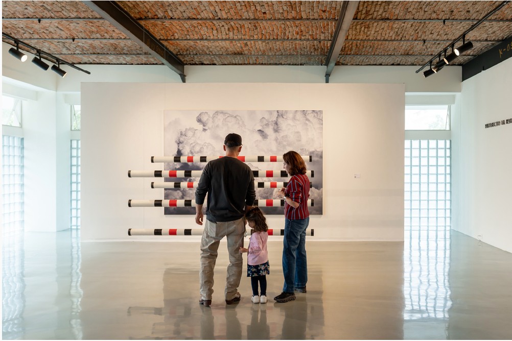 Un hombre, una mujer y una niña observan una pintura de nubes y una instalación de tubos en una sala blanca.